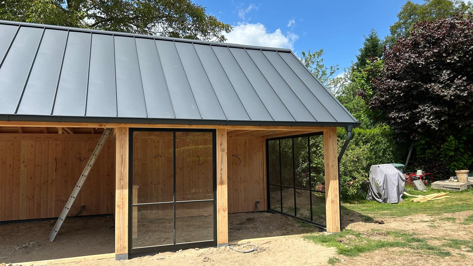 Houten buitenverblijf met open voorzijde, zwart potdekselwerk en een donker felsdak, omgeven door groene tuinplanten en bomen, gezien vanaf een stenen trap onder een deels bewolkte lucht.